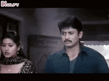 a man and a woman are standing next to each other in a room . the man has a mustache .