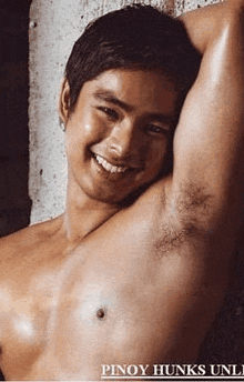 a shirtless man is leaning against a wall with his arm outstretched and smiling .