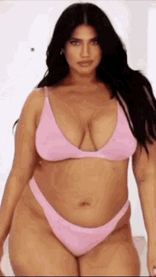 a plus size model is wearing a pink bikini and standing in front of a white wall .