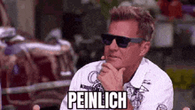 a man wearing sunglasses and a white shirt is sitting down with his hand on his chin and the word peinlich on his face .