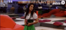 a woman in a green skirt is standing in a room with a milele logo on the bottom right