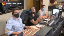 three men wearing face masks are sitting at a desk with eltrecetv.com written on the wall behind them