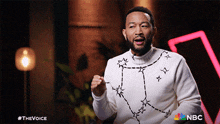 a man wearing a white sweater with stars on it is sitting in front of a nbc logo