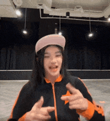 a girl wearing a pink hat and an orange jacket is pointing at the camera