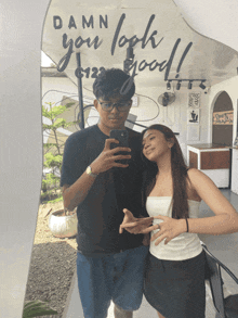 a man and a woman are taking a selfie in front of a sign that says damn you look good