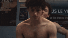 a shirtless man stands next to another shirtless man in front of a poster that says us le ve