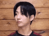 a young man with black hair is standing in front of a wooden wall and looking at the camera .