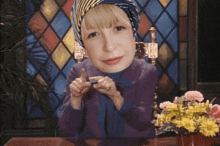 a little girl wearing a head scarf is sitting at a table with flowers in front of a stained glass window