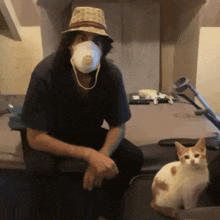 a man wearing a hat and a face mask sits next to a cat
