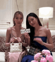 two women sitting on a couch holding up a sign that says bazaar