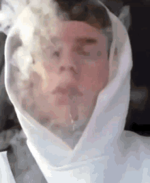 a man in a white hoodie is smoking a cigarette with smoke coming out of his face .