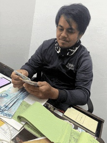 a man is sitting at a desk counting money and wearing a shirt that says ' criminal ' on the front