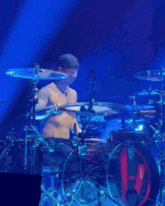 a shirtless man playing drums on a stage with a blue light behind him