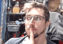 a man wearing glasses and a black shirt holds his finger to his mouth