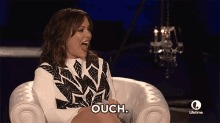 a woman is sitting in a chair with her mouth open and the word ouch is on the screen behind her