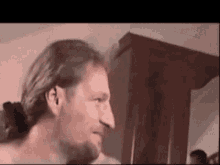 a man with long hair and a ponytail is standing in a room with a wooden cabinet in the background .