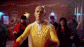 a bald man in a yellow costume is dancing in a room with other people .