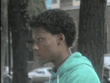 a man with curly hair is wearing a green hoodie and sitting under a tree .