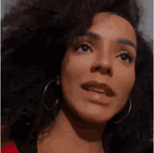a woman with curly hair and hoop earrings is wearing a red shirt and looking at the camera .