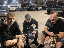 three men are sitting in a parking lot with one wearing a black sweatshirt with the letter f on the sleeve