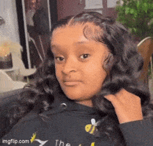 the girl is wearing a black hoodie with a bee on it and is looking at the camera .