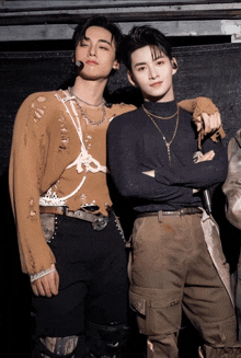 two men standing next to each other with their arms crossed and one wearing a brown sweater