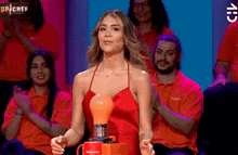 a woman in a red dress is holding a light bulb in front of a group of people applauding