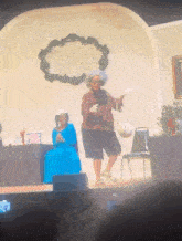 a woman in a blue dress is sitting in a chair while a woman in a hat is dancing on a stage