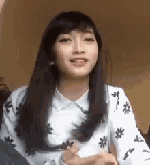 a woman with long black hair is wearing a white shirt with flowers on it and making a funny face .