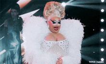 a drag queen in a white feathered outfit is smiling and holding a fan