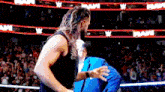 a man in a black tank top is standing in a wrestling ring with a crowd behind him .