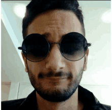 a man with a beard wearing round sunglasses looks at the camera