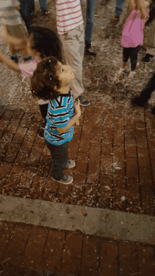 a young boy in a blue and white striped shirt stands on a brick sidewalk with his arms in the air