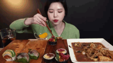 a woman in a green sweater is eating sushi with chopsticks on a wooden table