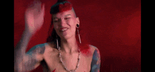 a woman with tattoos and red paint on her face is smiling and waving her hand .