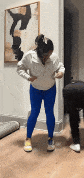 a woman in blue leggings and a white sweatshirt is standing on a wooden floor .