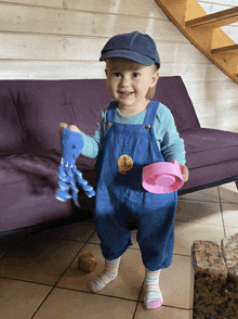 a little girl wearing overalls and a hat holds a stuffed animal