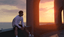 a man in a white sweatshirt sits on a ledge overlooking the ocean at sunset
