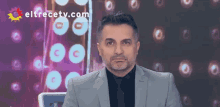 a man in a suit and tie is sitting in front of a screen that says eltrecetv.com on it