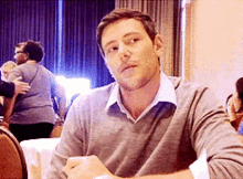 a man in a grey sweater is sitting at a table in a room .