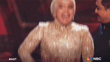 a woman in a gold dress is standing next to a man with the nbc logo on the bottom right