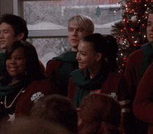 a group of people are singing in front of a christmas tree and wearing scarves with the letter t on them