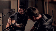 two men playing guitars in front of microphones in a room