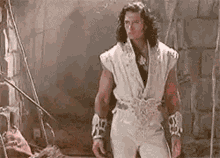 a man with long hair is standing in front of a brick wall in a white outfit .