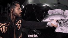 a man in a black and gold jacket holds a dead fish in front of a car