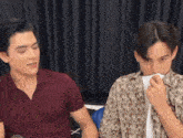 two men are sitting next to each other in front of a microphone and one of them is blowing his nose .