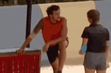 a man in a red tank top is sitting on a box next to a woman in a black shirt .