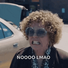 a woman with curly hair is wearing sunglasses and making a funny face while standing in front of a car .