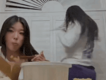 a woman is sitting at a table eating noodles with chopsticks while another woman looks on .