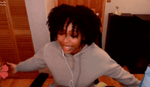 a woman wearing headphones and a gray hoodie smiles with her arms outstretched